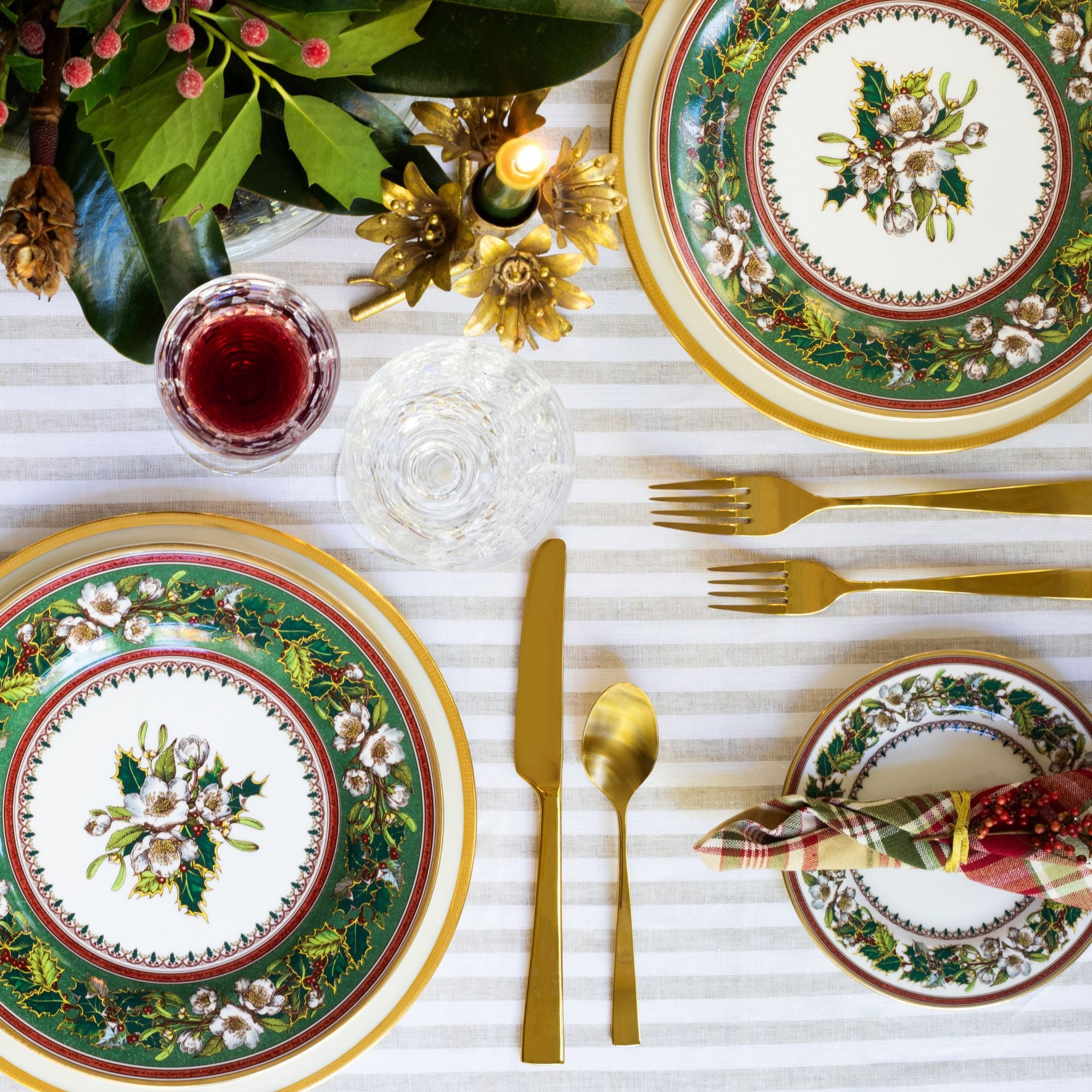 Holiday place setting with dinner plates featuring holly roses, gold luster flatware, and crystal.